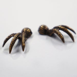 May include: Two bronze-coloured claw-shaped objects are shown against a white background. Each claw has three sharply curved talons extending from a textured base. The objects appear to be jewellery components or decorative elements.