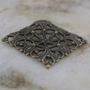 May include: A square-shaped metal charm with a decorative, intricate design. The charm is made of a dark, antique-looking metal and has a filigree pattern.