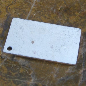 May include: A silver rectangular tag with a small hole at the top left corner. The tag has a slightly textured surface and a few small dark spots.