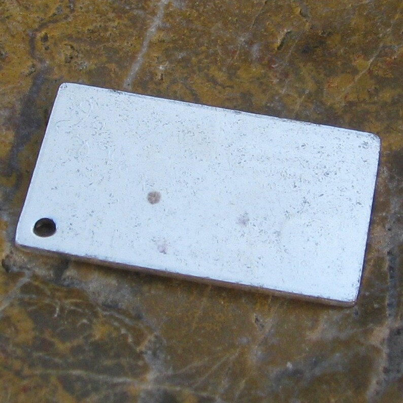 May include: A silver rectangular tag with a small hole at the top left corner. The tag has a slightly textured surface and a few small dark spots.