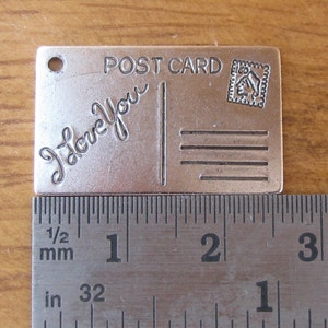 May include: Silver metal charm shaped like a postcard with the text "POST CARD" and "I love you" in a cursive font. The charm has a hole at the top for hanging.