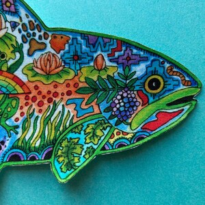 May include: A colorful fish-shaped patch with a green outline. The fish is covered in a variety of patterns and designs, including flowers, leaves, and geometric shapes. The fish's eye is black with a yellow ring around it.