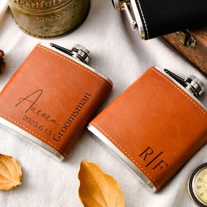 Könnte beinhalten: Zwei braune Lederflaschen mit silbernen Akzenten. Eine Flasche ist mit "Aurora 2025.6.13 Groomsman" graviert, die andere mit "R/F". Eine Vintage-Taschenuhr und andere dekorative Elemente sind ebenfalls vorhanden.