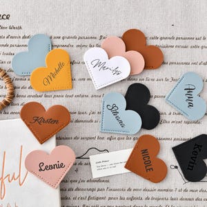 Peut inclure: Un ensemble de dix marque-pages en forme de cœur avec des noms différents écrits dessus. Les marque-pages sont en cuir et sont disponibles dans une variété de couleurs, notamment le rose, le bleu, le marron et le noir.
