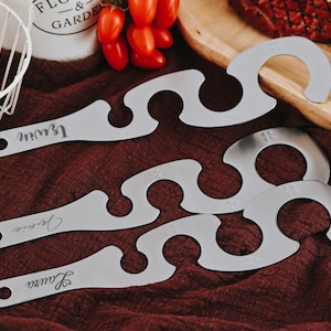 Könnte beinhalten: Vier silberne Grillwerkzeuge mit wellenförmigem Design und dem eingravierten Wort "Leanne" auf jedem. Die Werkzeuge befinden sich auf einer roten Stoffoberfläche.