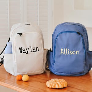 May include: Two personalized canvas backpacks, one white with black text "Waylan" and one blue with yellow text "Allison".