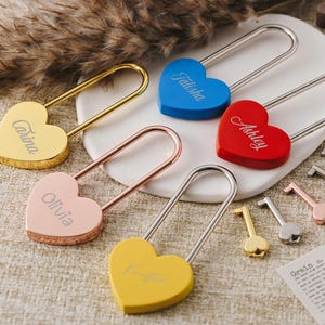 Può includere: Cinque lucchetti a forma di cuore con nomi personalizzati: Talitha, Ashley, Carina, Olivia e Katia. I lucchetti sono di vari colori: blu, rosso, oro, rosa e giallo. I lucchetti sono su una superficie marrone chiaro con diverse chiavi.