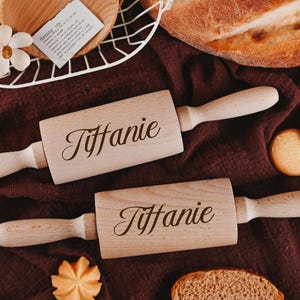 Pu&ograve; includere: Due mattarelli di legno con il nome "Tiffanie" inciso su di essi. I mattarelli sono su una superficie di tessuto marrone con alcuni biscotti e pane sullo sfondo.