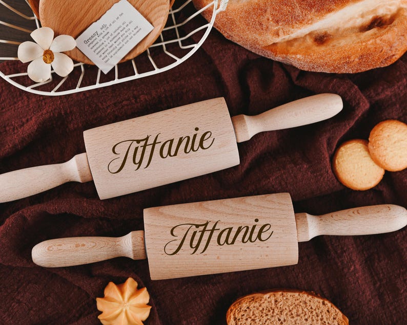 Pu&ograve; includere: Due mattarelli di legno con il nome "Tiffanie" inciso su di essi. I mattarelli sono su una superficie di tessuto marrone con alcuni biscotti e pane sullo sfondo.
