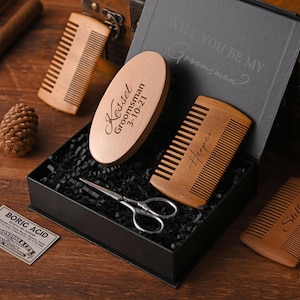 May include: A wooden groomsmen gift set in a black box. The set includes a comb, a beard brush with the name "Kessel" and the date "3-10-21", a pair of scissors, and a small label that says "Boric Acid". The box is lined with black paper.