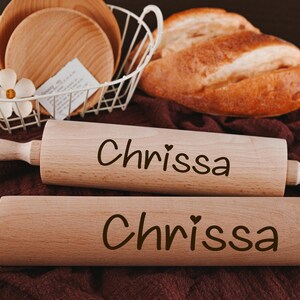 Pu&ograve; includere: Due mattarelli di legno con il nome "Chrissa" inciso su di essi. I mattarelli sono su una superficie marrone.