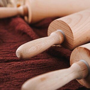 Pu&ograve; includere: Tre mattarelli di legno su una superficie di stoffa marrone. I mattarelli sono in legno chiaro e hanno una finitura liscia.