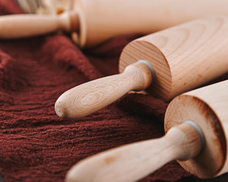 Pu&ograve; includere: Tre mattarelli di legno su una superficie di stoffa marrone. I mattarelli sono in legno chiaro e hanno una finitura liscia.