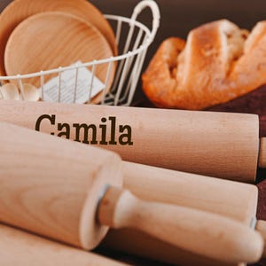 Pu&ograve; includere: Tre mattarelli di legno, uno con il nome "Camila" inciso su di esso. I mattarelli sono su una superficie marrone con un panno marrone e bianco sullo sfondo.