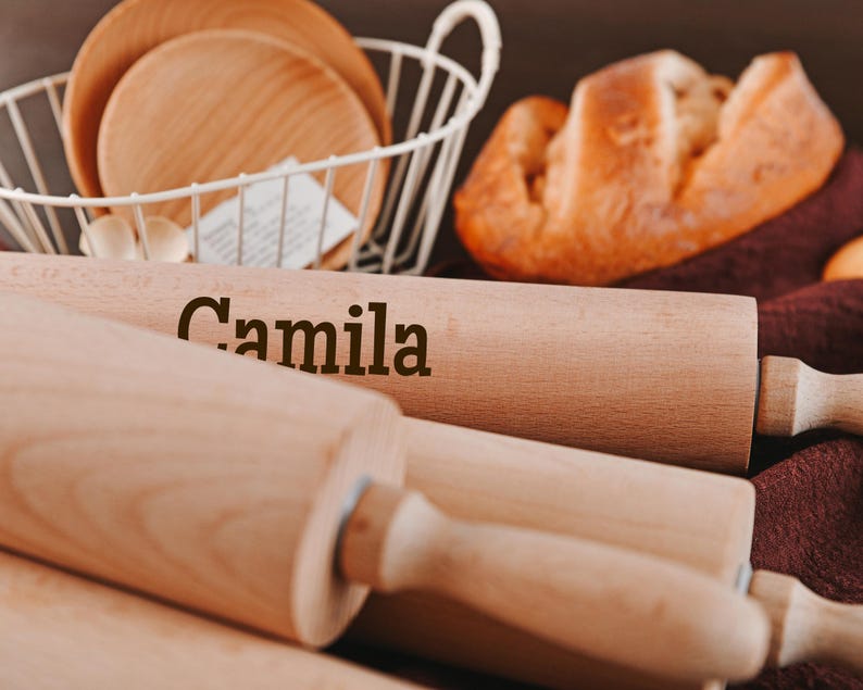 Pu&ograve; includere: Tre mattarelli di legno, uno con il nome "Camila" inciso su di esso. I mattarelli sono su una superficie marrone con un panno marrone e bianco sullo sfondo.