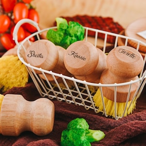 Puede incluir: Cuatro cepillos de fregar de madera con cerdas amarillas en una cesta de alambre blanca. Los cepillos tienen los nombres "Shelia", "Kassie" y "Turelle" grabados en ellos.