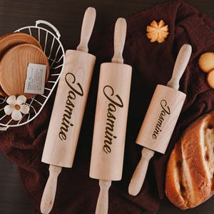Pu&ograve; includere: Tre mattarelli di legno con il nome "Jasmine" inciso su ciascuno. I mattarelli sono su una superficie di stoffa marrone con un pane e biscotti.