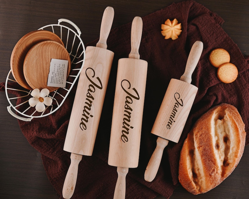 Pu&ograve; includere: Tre mattarelli di legno con il nome "Jasmine" inciso su ciascuno. I mattarelli sono su una superficie di stoffa marrone con un pane e biscotti.