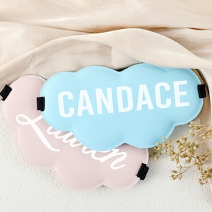 Op de afbeelding: Twee wolk-vormige slaapmaskers. Eén is lichtblauw met de naam "CANDACE" in wit, en de andere is roze met de naam "Lauren" in witte letters. Beide hebben zwarte elastische bandjes.