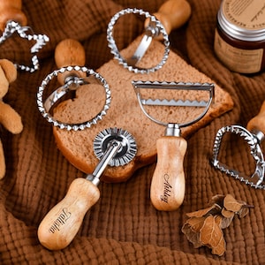 Puede incluir: Una colección de utensilios para hornear con mangos de madera y bordes de corte de acero inoxidable, que incluyen un cortador en forma de estrella, un cortador en forma de corazón y un cortador de ravioles. También hay un pequeño oso de peluche y un tarro de vela.