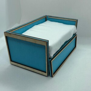 Puede incluir: Una cama en miniatura con un marco azul claro y una sábana blanca. El marco de la cama tiene un borde de madera natural. La cama está diseñada para una casa de muñecas u otro entorno en miniatura.