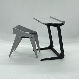 Puede incluir: Un juego de silla y mesa en miniatura de color gris. La silla tiene un asiento triangular y patas anguladas. La mesa tiene una parte superior rectangular sostenida por una base negra y curvada. El conjunto está diseñado con una estética moderna y minimalista.