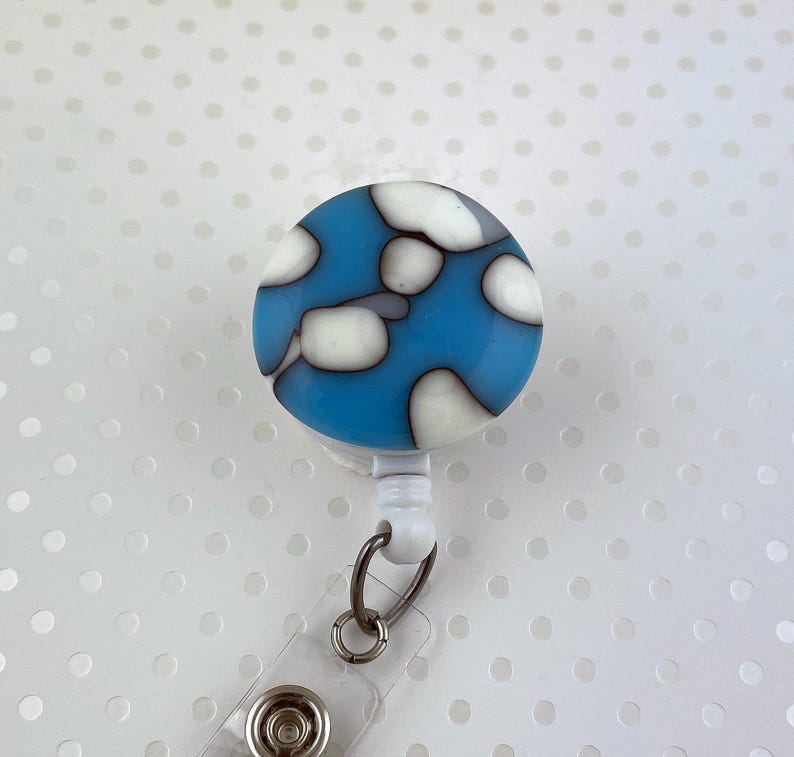 May include: A round, blue and white badge reel with a cloud-like pattern. The reel has a clear plastic strap with a metal clip and a button snap. The design is a mix of light blue, white, and gray.