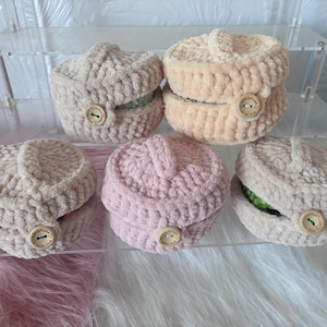 May include: Five round, crocheted containers made of soft, plush yarn in shades of beige, peach, and light pink. Each container features a lid with a small handle and a wooden button on the side engraved with the text Handmade with love.