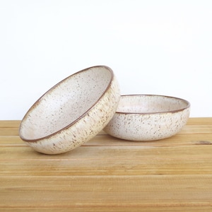 May include: Two round ceramic bowls with a speckled cream and off-white glaze. The bowls feature a dark brown rim and a textured finish with small brown flecks distributed across the interior and exterior surfaces of the rounded vessels.