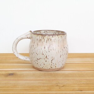 May include: A handmade ceramic mug with a speckled, off-white glaze and a brown rim. The mug has a large, curved handle and sits on a wooden surface. The mug is approximately 4 inches tall.