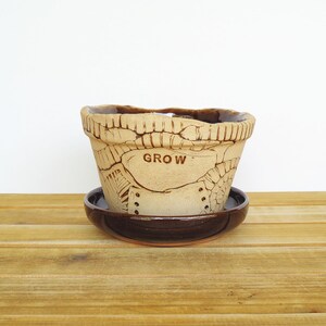 May include: A ceramic planter with a brown glaze and a distressed finish. The planter has a saucer and the word "GROW" is written on the side.