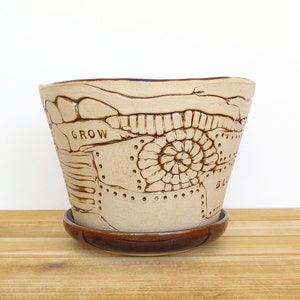 May include: A beige ceramic planter with a brown glaze and a decorative design. The planter has a saucer and the word "GROW" is embossed on the side.
