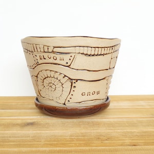 May include: A beige ceramic planter with a brown glaze and a decorative design. The planter has the words "Bloom" and "Grow" inscribed on it.