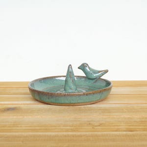 May include: A round ceramic ring dish with a glossy teal and brown glaze. The dish features a central conical post for holding rings and a small sculpted bird figure perched on the inner rim, both finished in the same teal and brown glaze.
