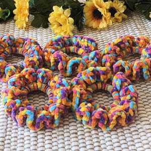 May include: Six colorful crocheted hair scrunchies in shades of purple, orange, yellow, and blue. The scrunchies are arranged in a circle on a white textured surface.