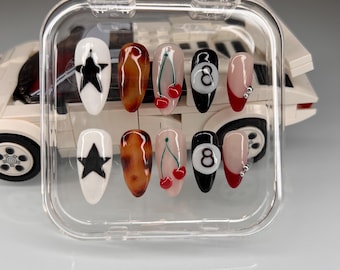 May include: A set of ten false nails in a clear plastic container. The nails are decorated with various designs, including a black star, a brown and black tortoise shell pattern, a red cherry, and a black and white 8-ball.