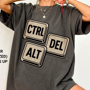 May include: Charcoal grey oversized t-shirt with the words "CTRL", "ALT", and "DEL" in a keyboard key design. The shirt has a relaxed fit and is made of a soft material. The text "PEPPER (OVERSIZED) 1-2 SIZES UP" is printed on the left side.