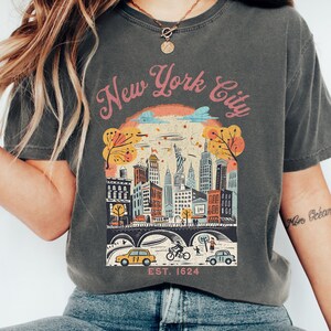 May include: Dark grey t-shirt with a colourful graphic of the New York City skyline, featuring buildings, trees, and vehicles. The text "New York City" is arched above the illustration, with "EST. 1624" below. The shirt is a Pepper colour.