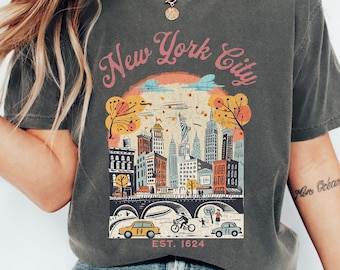 May include: A charcoal gray short-sleeve t-shirt featuring a graphic print of the New York City skyline with the text New York City and EST. 1624. The illustration includes buildings, trees, a bridge, cars, and a cyclist in orange, blue, and cream tones.