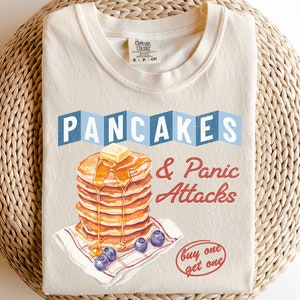 May include: Ivory t-shirt featuring a graphic of a stack of pancakes with syrup and butter, and the text "Pancakes & Panic Attacks" in red and blue. The text "buy one get one" is also included in red.