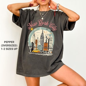 May include: An oversized charcoal grey t-shirt featuring a vintage-style graphic of the New York City skyline. The graphic includes a yellow taxi cab, the Empire State Building, and the Statue of Liberty. The text "New York City" is printed in pink lettering.