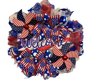 May include: A patriotic wreath with red, white, and blue mesh, bows, and pinwheel accents. The wreath features a sign that says "Welcome" in white script. The design incorporates stars and stripes, perfect for celebrating Independence Day.