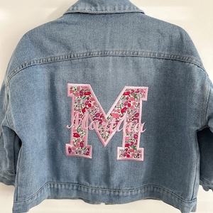 May include: A blue denim jacket with a large pink floral letter M on the back. The letter is outlined in green and has the name "Martina" embroidered in pink below it.