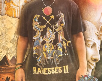 Puede incluir: Camiseta negra con un estampado gráfico de un carro tirado por dos caballos. El carro es conducido por un faraón con una corona y un bastón. El texto "RAMESES II" está impreso debajo de la imagen. La camiseta también tiene un pequeño logotipo con el texto "SUPERIOR WARRIOR".