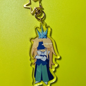 May include: A clear acrylic keychain featuring a cartoon character with long blonde hair, a blue crown, and a blue and teal outfit. The keychain has a gold star-shaped clasp.