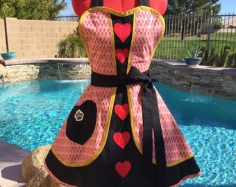 May include: A red and white patterned apron with black and gold trim, featuring red heart accents and a black pocket with a crown emblem. The apron has a black waist tie and halter straps, displayed on a red dress form.