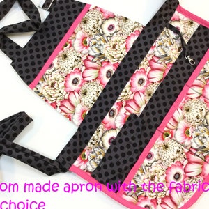 May include: Custom-made apron featuring a floral pattern with pink and white flowers, accented by black polka dot fabric and pink trim. The apron has a long strap and a metal clasp.