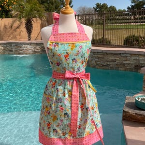 May include: A colorful apron with a floral and cocktail print. The apron features a pink bow, pink trim, and a light blue background. The apron is displayed on a mannequin near a swimming pool.