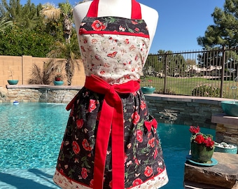 May include: A floral apron with a red bow and straps. The apron features a black skirt with red roses and a cream-colored bodice with butterflies. The apron is displayed on a mannequin in front of a pool.
