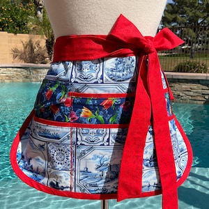 May include: A half apron with a red waist tie and trim. The apron features a blue and white toile pattern with red and yellow floral accents. The apron is displayed on a mannequin.